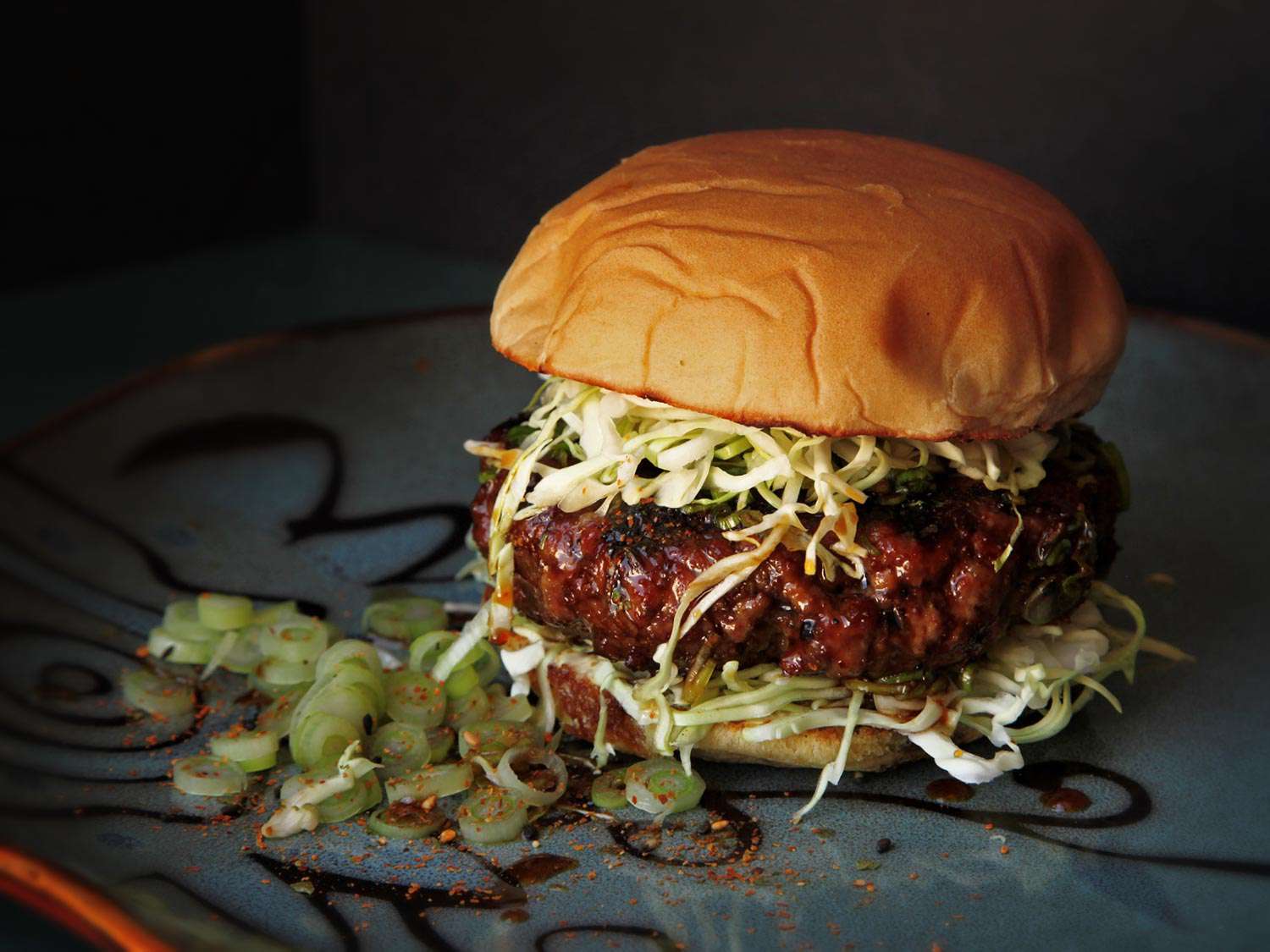 A teriyaki burger, served on a painted ceramic plate, emerging from the shadows.
