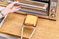 A hand holding the door of the Breville the Compact Smart Oven next to tongs holding cheese toast