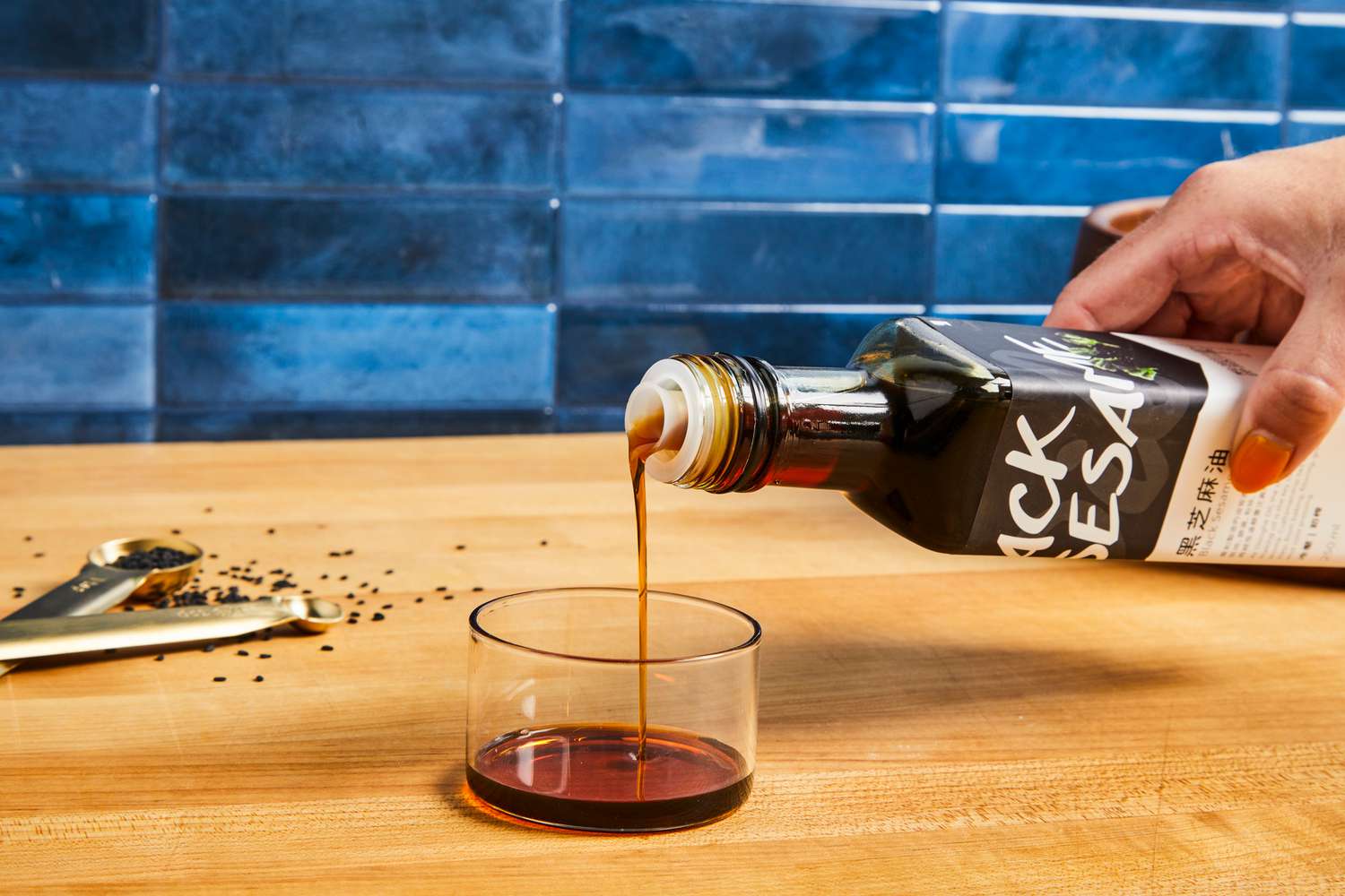 Pouring sesame oil into a small glass container, measuring spoons and sesame seeds nearby on a wooden surface