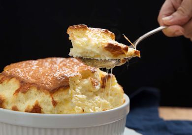 A large savory cheese souffle with someone holding a spoon of souffle above the casserole. 