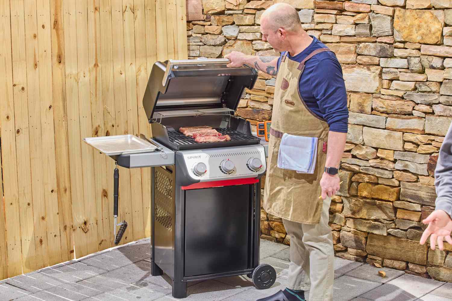 A person holding the lid handle of the Weber Spirit E-310 Gas Grill while looking at raw steaks on the grill