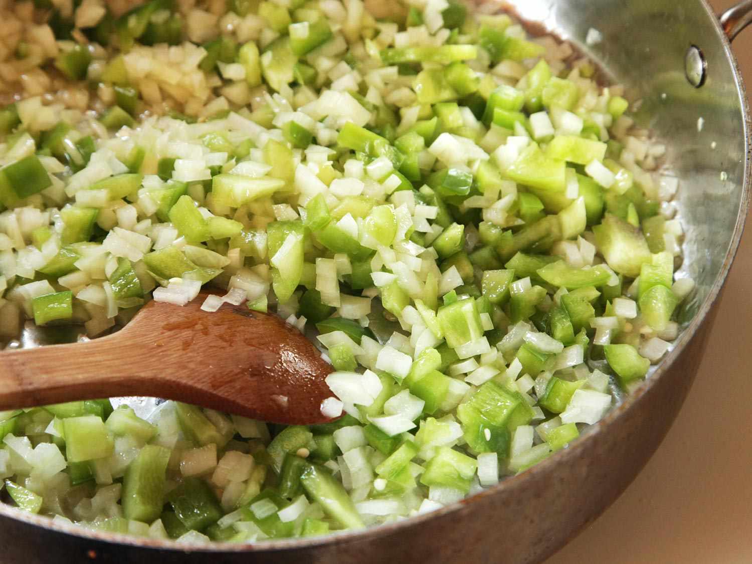 Diced onion and green pepper cooking in the sauté pan with butter and olive oil. A wooden spoon is stirring the mixture.