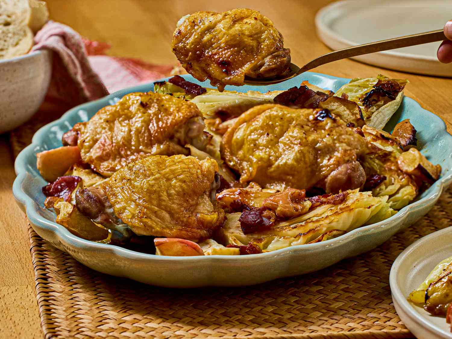 A dish of roasted chicken thighs with bacon apple slices and vegetables on a plate presented on a woven surface
