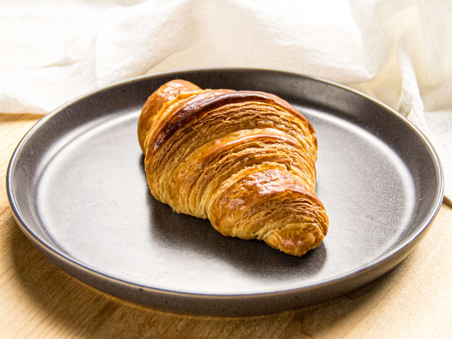 A croissant on a round plate displayed on a wooden surface with a lightcolored cloth in the background