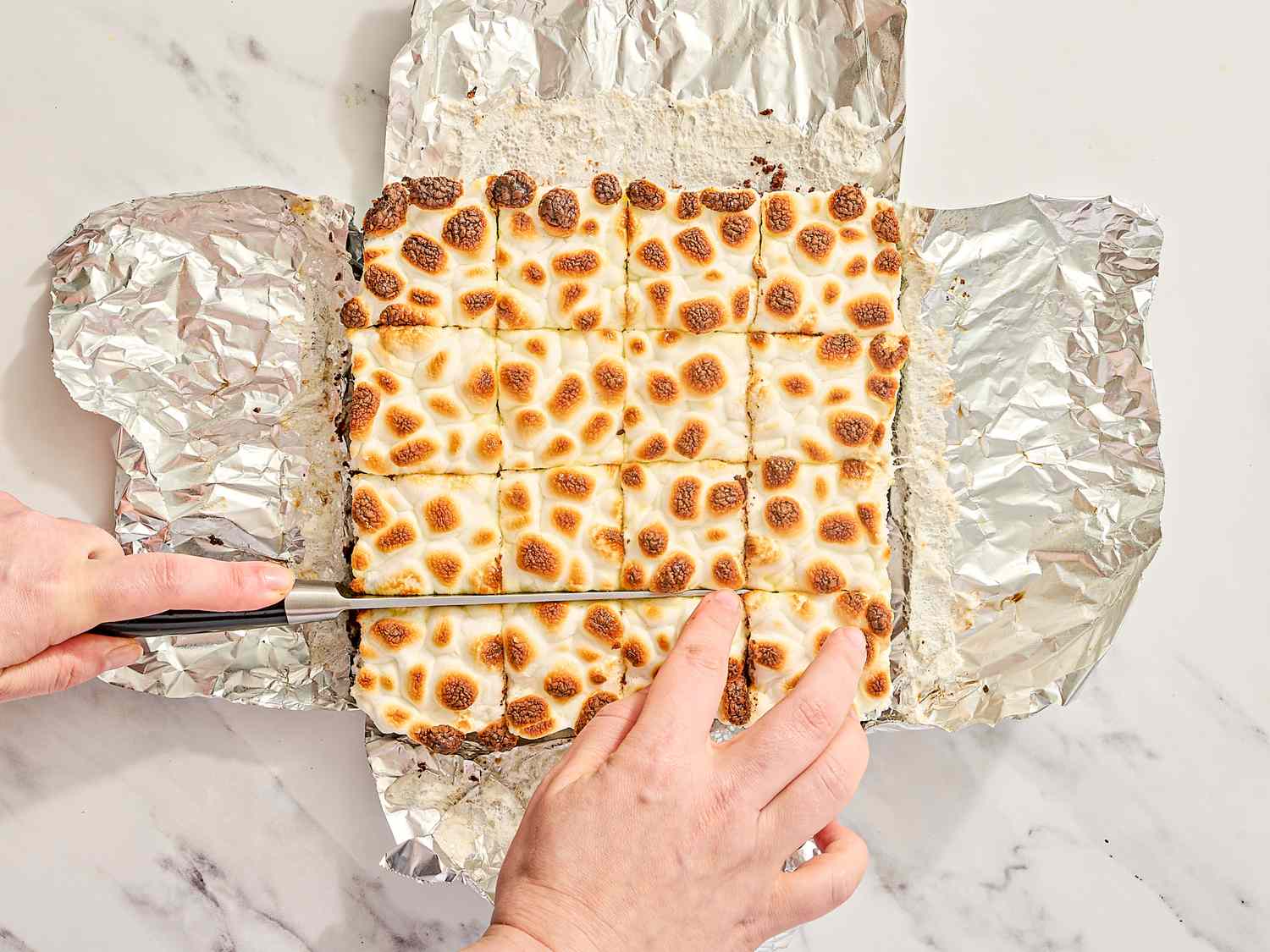 Hands slicing a tray of toasted marshmallowcovered brownies on foil