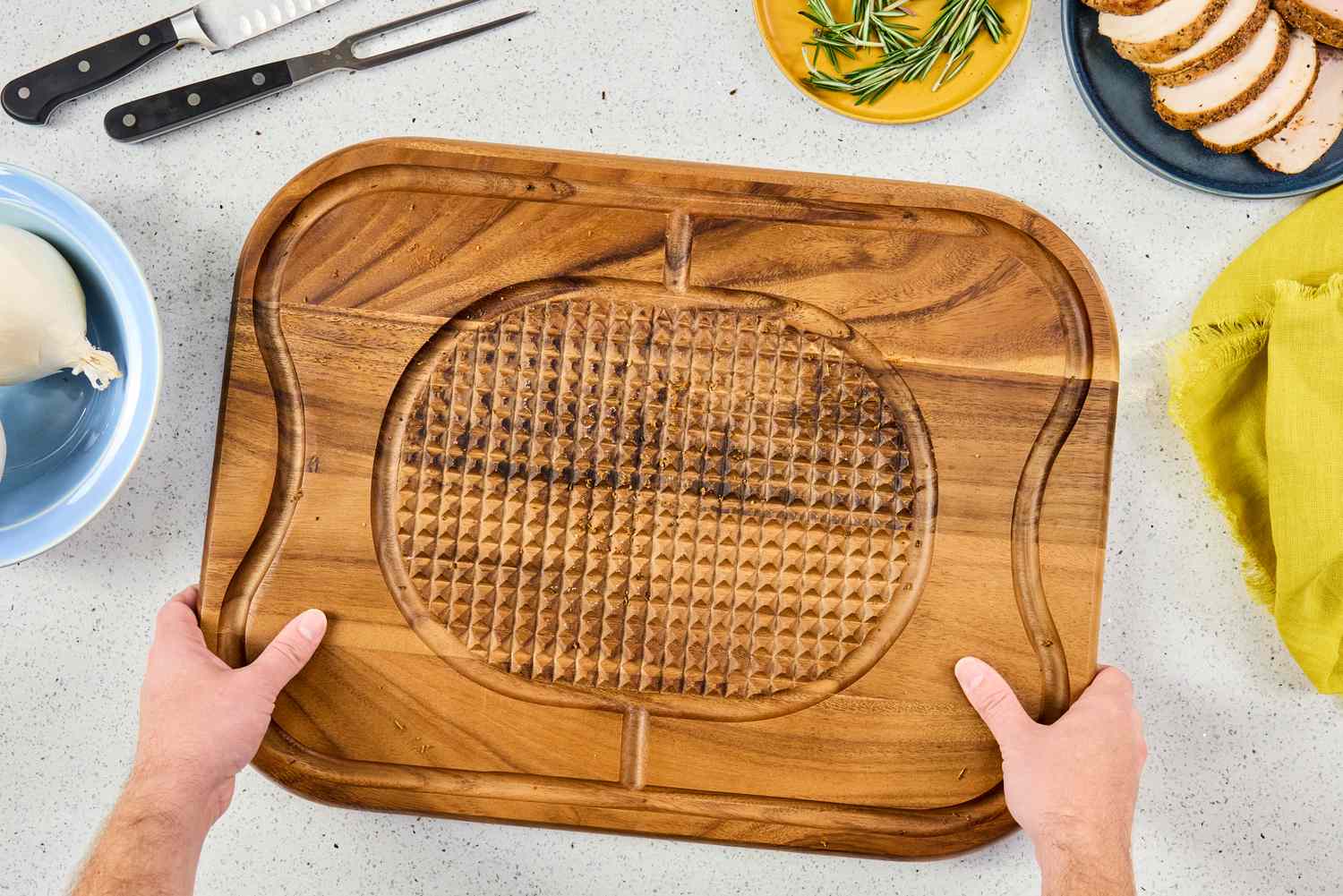 Hands holding a Lipper International Acacia Cutting Board with Grid Grip on a light countertop with utensils and food ingredients nearby