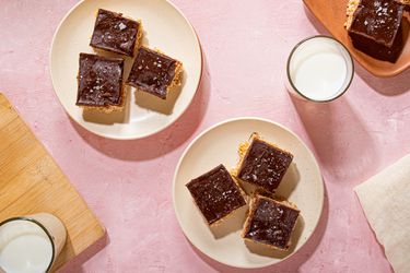 Plates of scotcheroos, chocolate-topped dessert bars, placed on a pink surface beside glasses of milk