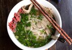 A bowl of pho with meat, noodles, herbs, and a pair of chopsticks resting on the side of the bowl