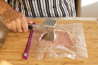 A meat tenderizer being used on a piece of meat in a plastic bag on a wooden cutting board