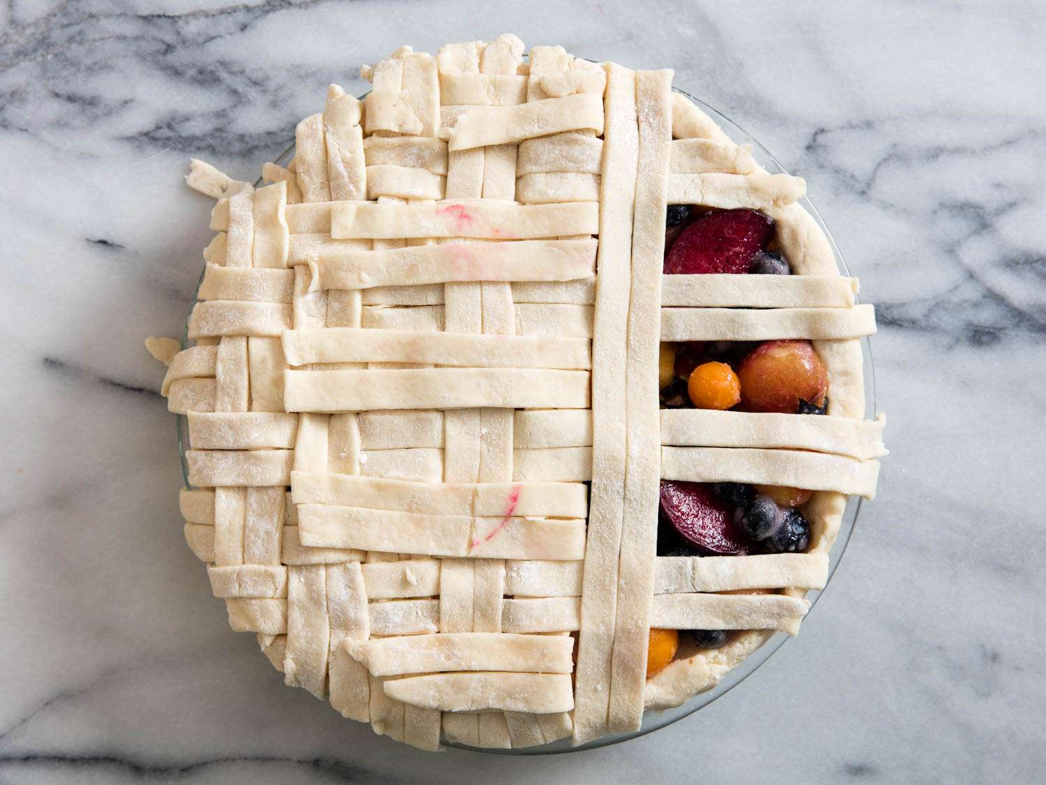 The basketweave top crust nearly assembled on top of the pie dish. Several gaps remain and the summer fruit filling is partially visible.