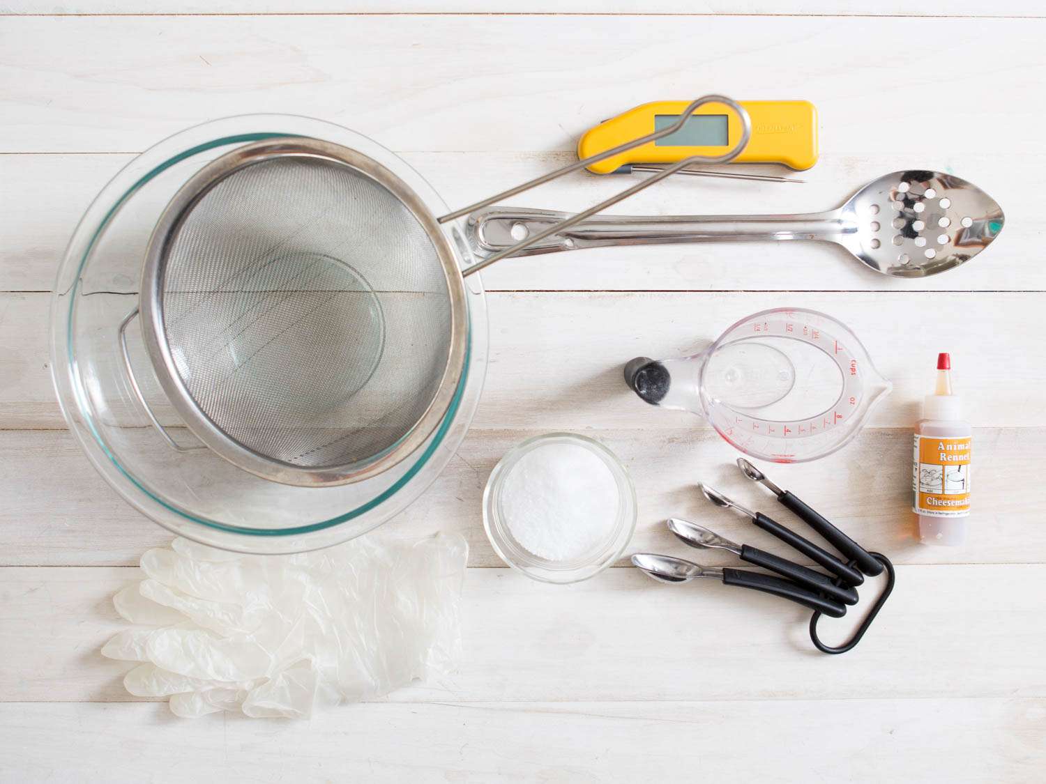 Supplies needed for making fresh mozzarella at home: bowl, strainer, thermometer, slotted spoon, measuring cup, measuring spoons, rennet, citric acid, and gloves.