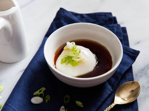 An onsen egg served at the table, cradled in a white bowl, half-submerged in soy-seasoned dashi. Sliced scallion is sprinkled over the top.