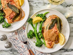 Two plates of chicken saltimbocca with sauce broccolini and lemon wedges on a table setting