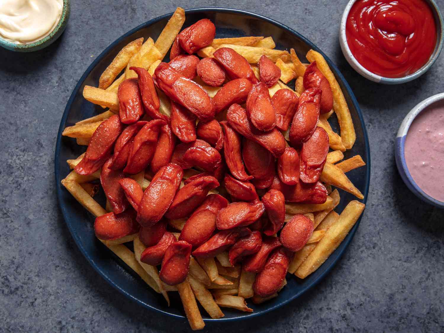 A platter of salchipapa shows fried sliced hotdogs and thick-cut French fries on a platter; a few sauces can be seen in dishes on the side for dipping.