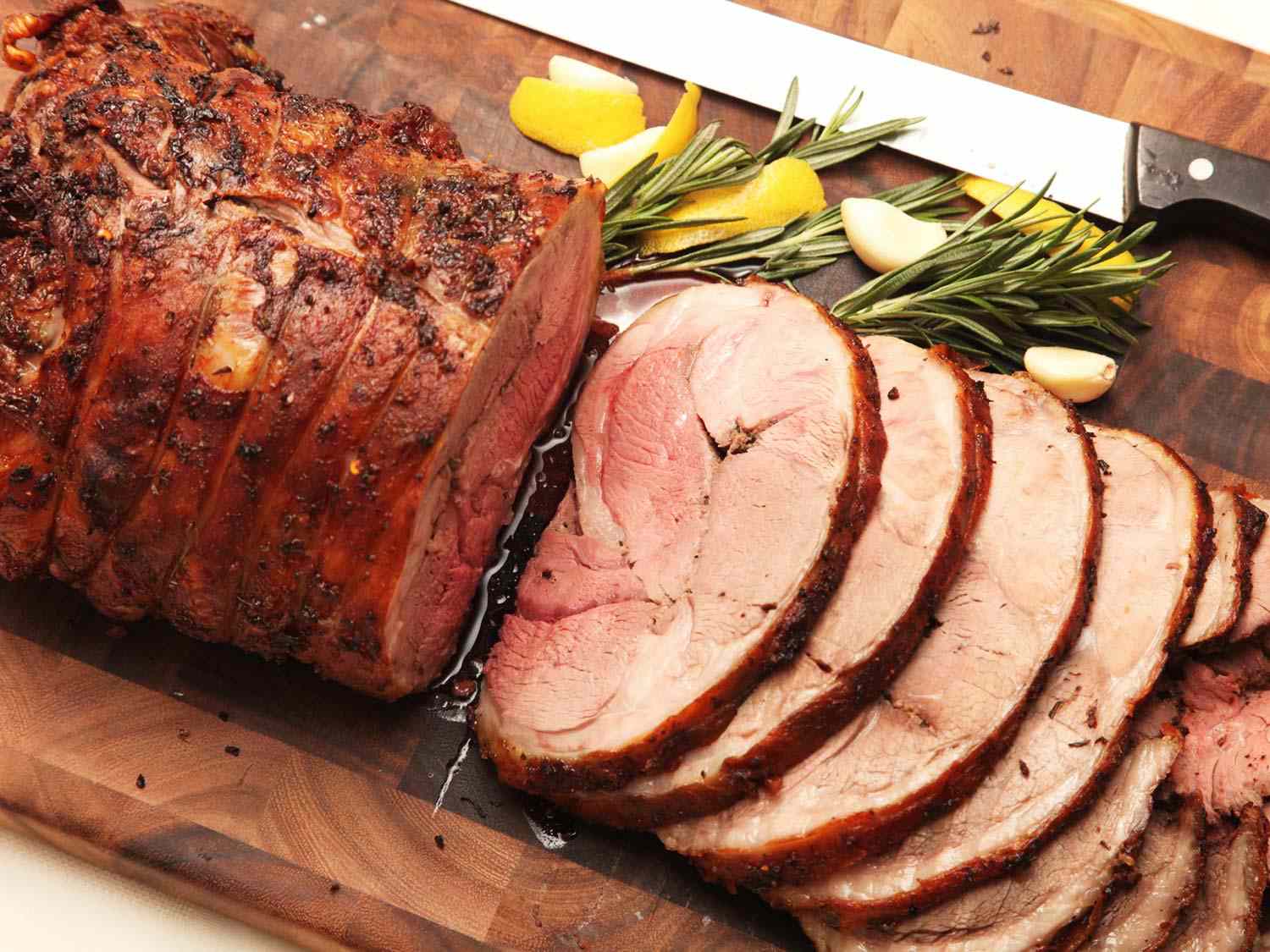 A slow-roasted leg of lamb sliced on a cutting board, flanked by rosemary sprigs, peeled garlic cloves, and wedges of lemon.