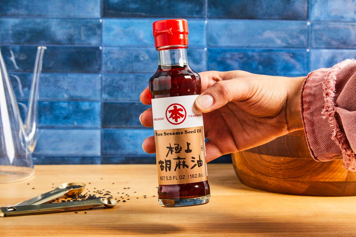 A hand holding a bottle of pure sesame seed oil labeled with Japanese text, on a wooden countertop with teaspoons and black sesame seeds