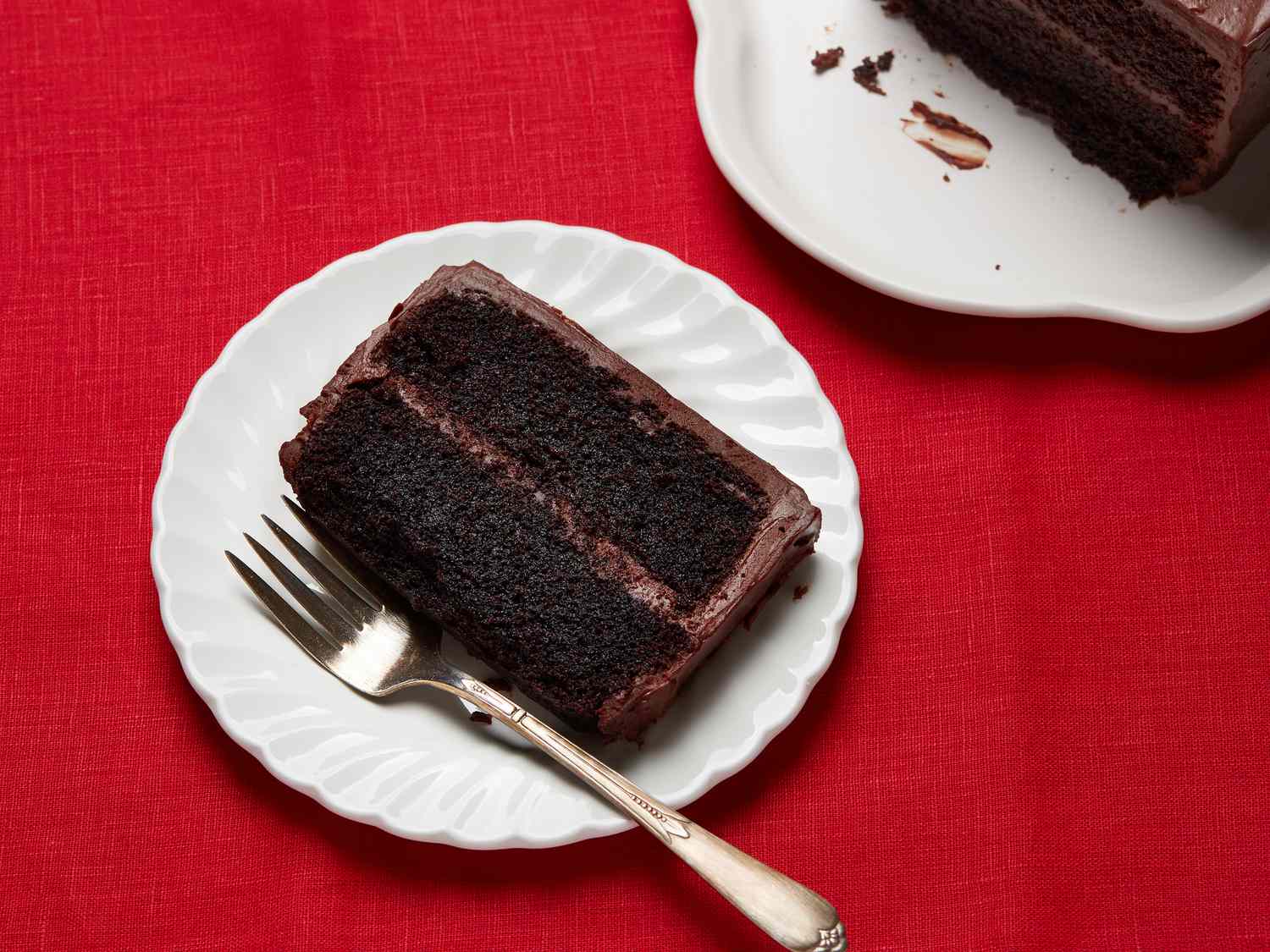 slice of cake on white plate, with cake to the side on white platter on a red fabric surface. 