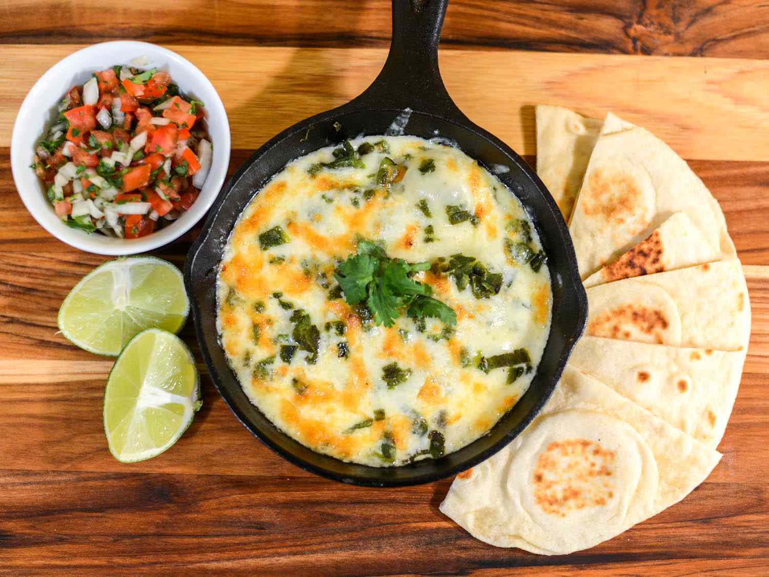 A skillet of queso fundido with flour tortillas, pico de gallo, and lime wedges