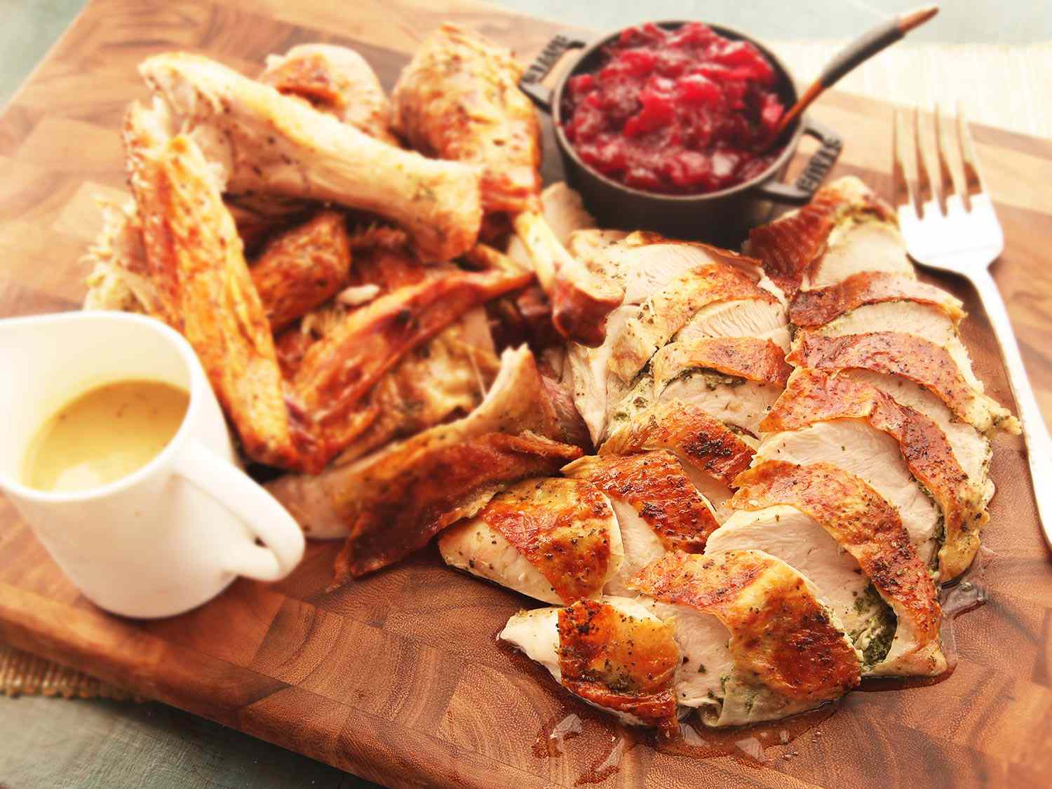 Cut up roasted turkey on a wooden cutting board with a pitcher of gravy and a bowl of cranberry sauce.
