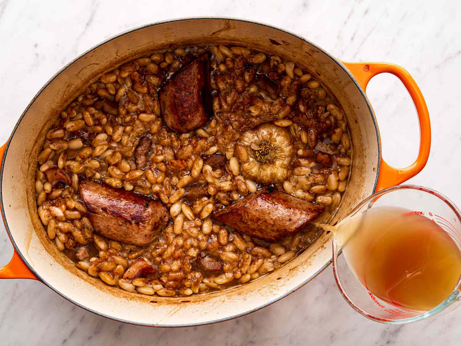 Pouring stock into pot filled with sausages and beans.