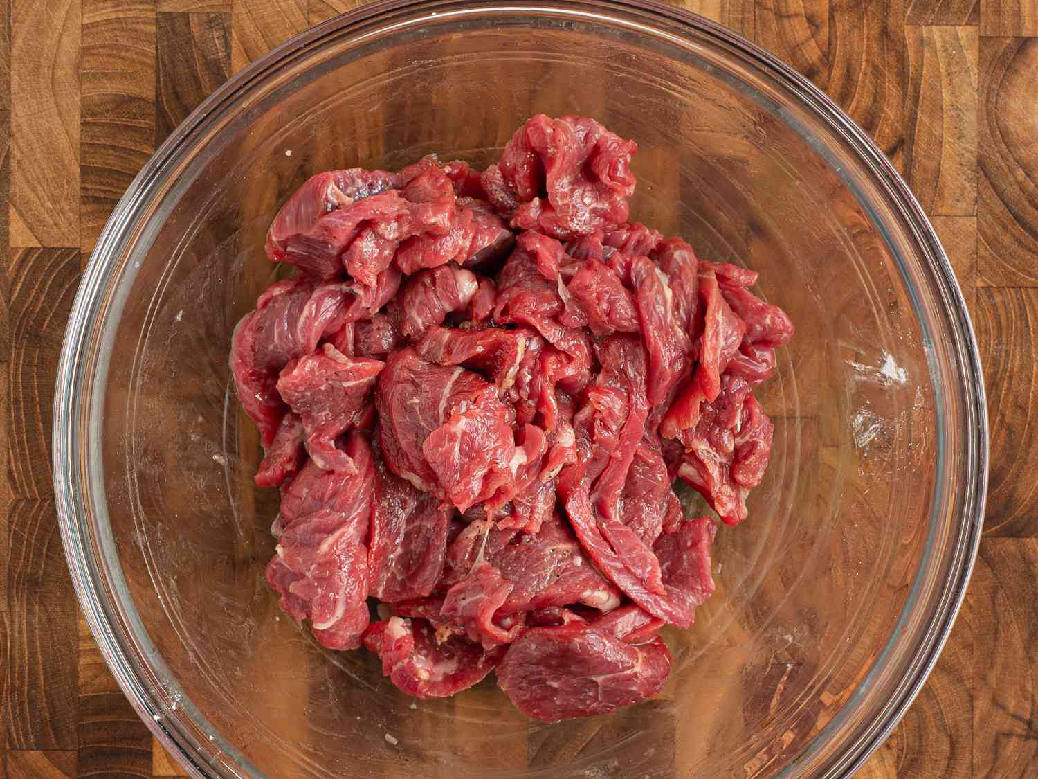 A glass bowl holding beef tossed with salt, sugar, pepper, wine, soy sauce, oil, and cornstarch. The bowl is on a wooden cutting board.