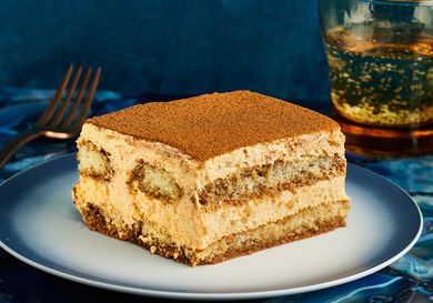 Slice of PSL Tiramisu on a stylish blue and white plate, with a blue tablecloth, sparkling water, and a gold fork