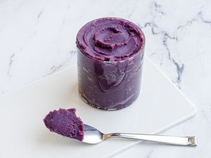 A glass jar of ube halaya placed on a white marble surface. A morsel of the stiff jam is perched on a spoon in front of the jar.
