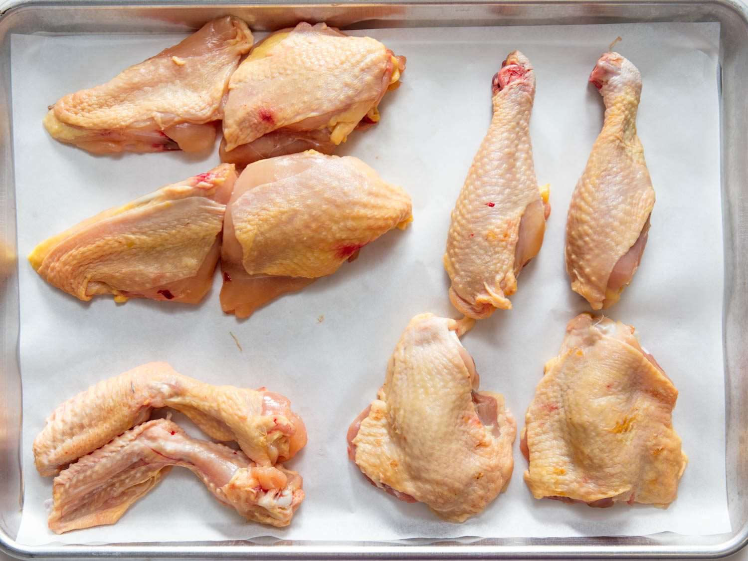 Various raw chicken parts (breasts, drumsticks, wings, and thighs) arranged on a parchment-lined metal sheet pan