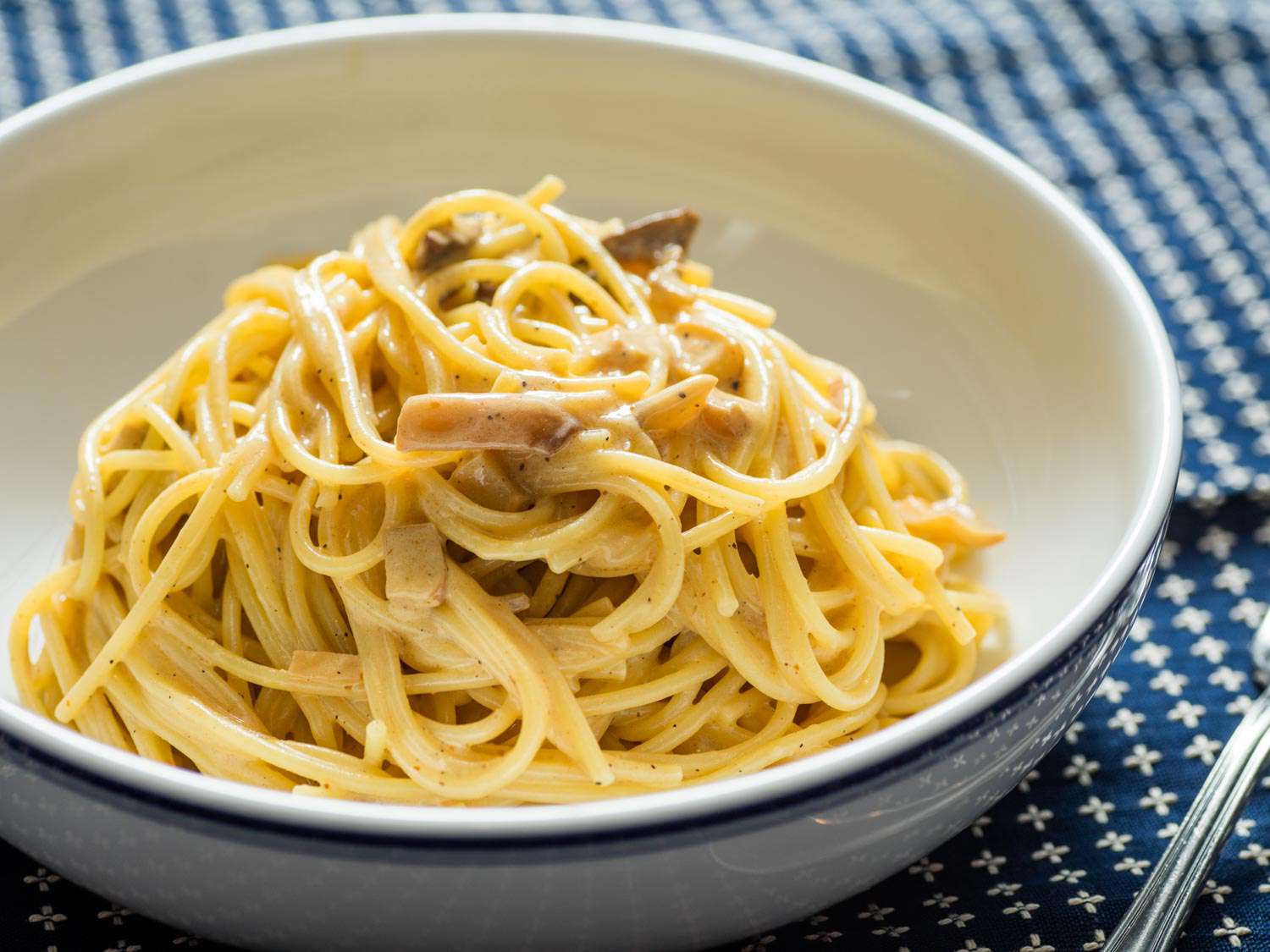 A white bowl of vegan carbonara is set on a blue, patterned table cloth.