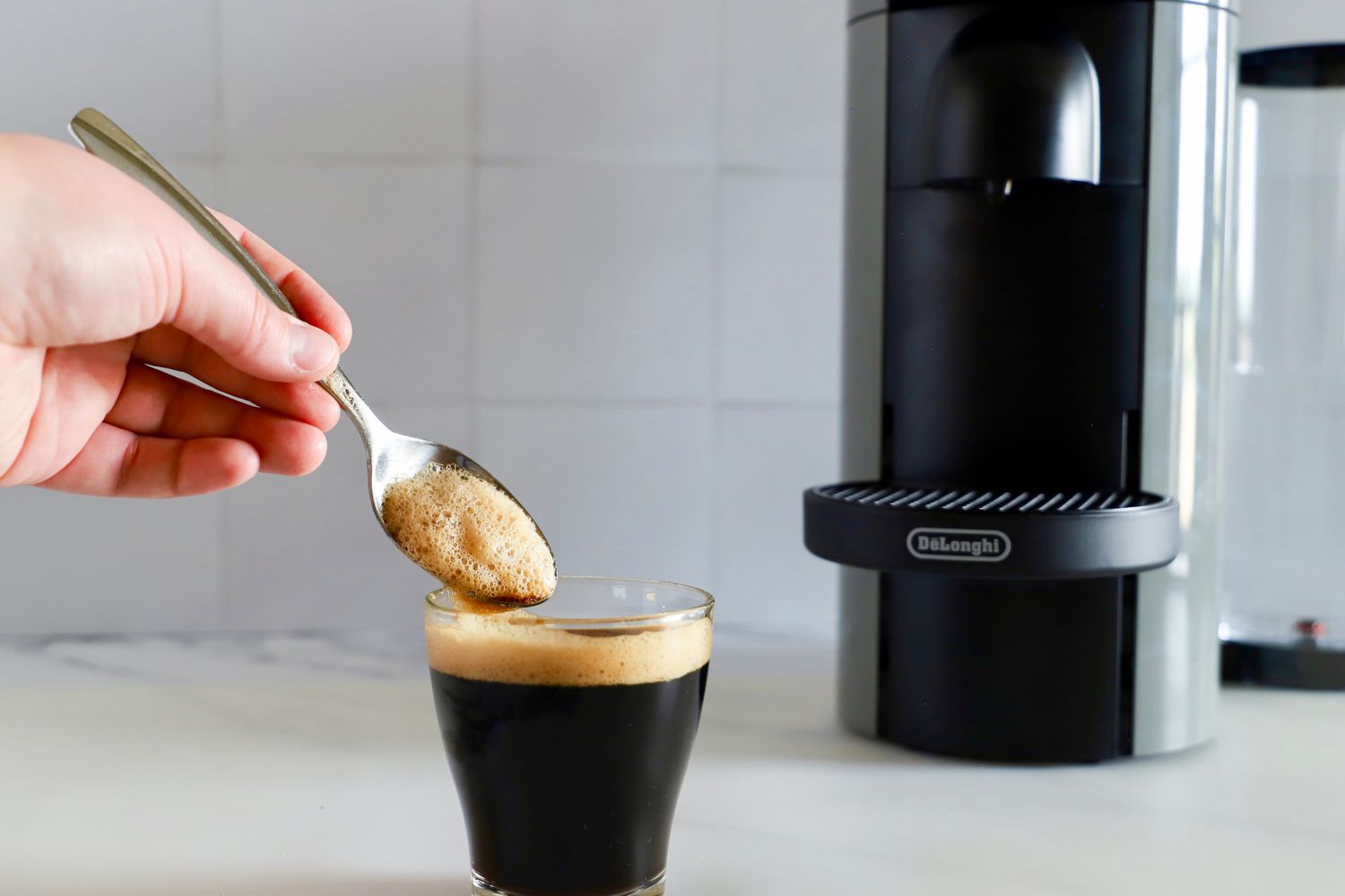 A hand holding a spoon in a cup of espresso next to the Nespresso VertuoPlus Coffee and Espresso Machine by De'Longhi