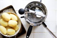 A food mill with boiled potatoes in a steamer basket on a white tile surface