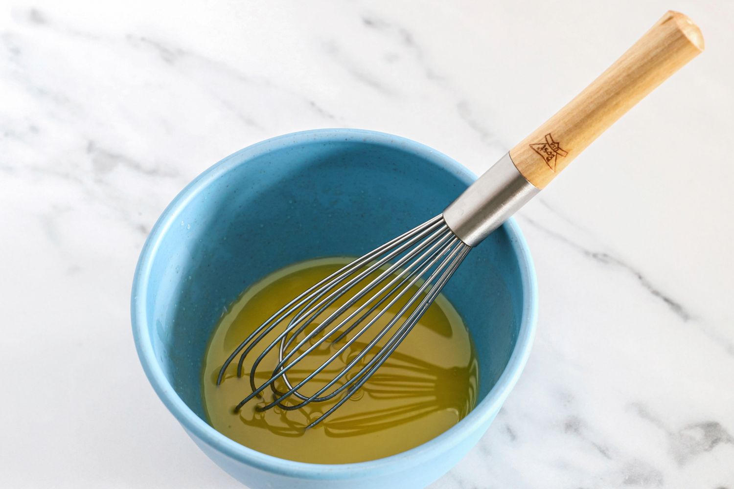 A whisk in a blue Bamboozle bowl containing a liquid mixture on a marble surface