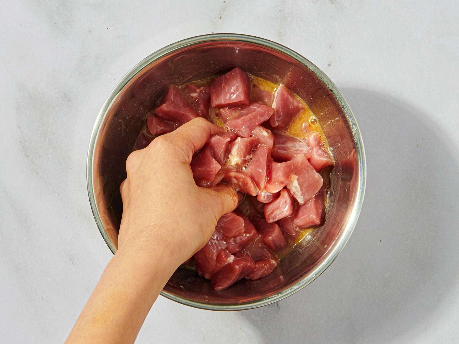 Hand mixing diced meat in a metal bowl containing marinade