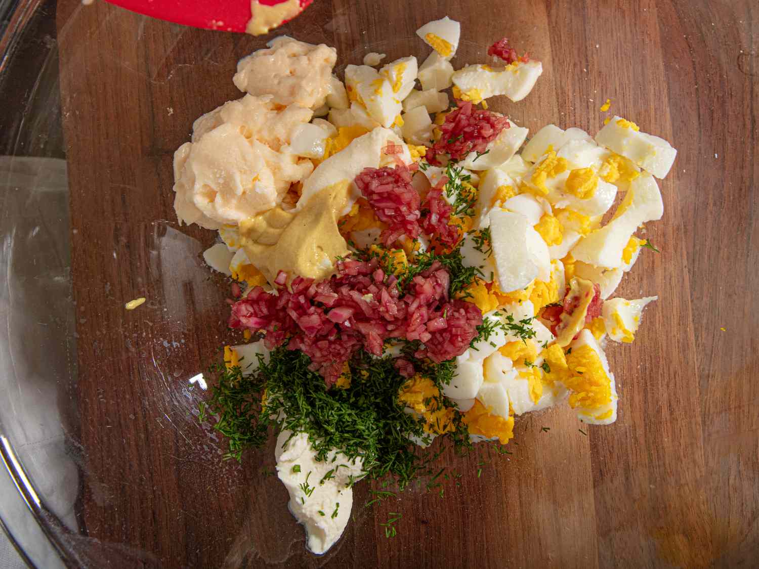 Ingredients for egg salad on a wooden surface, including diced boiled eggs, mayonnaise, mustard, and dill and pickled red onions