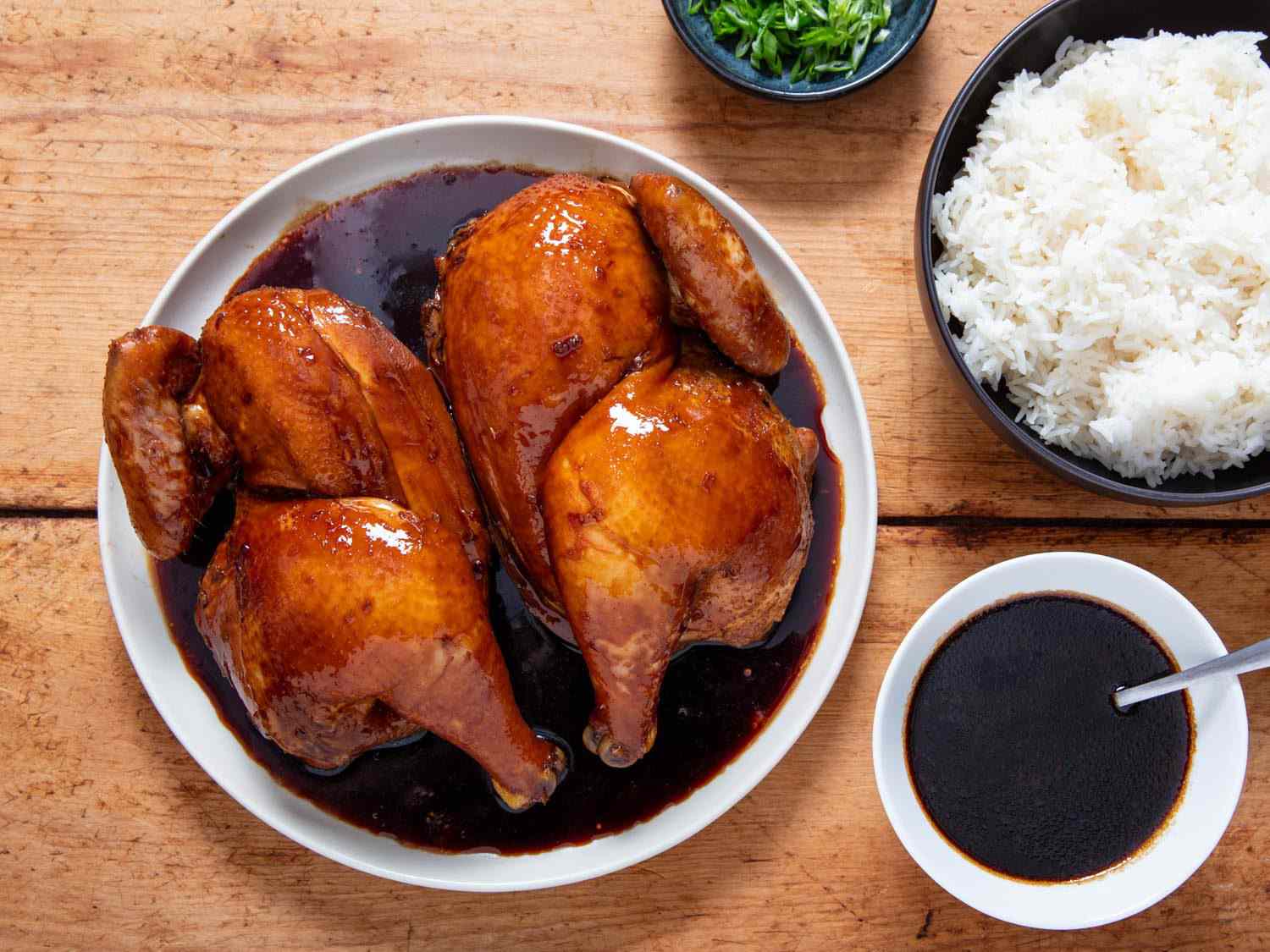 Overhead of soy sauce chicken with cola on a serving platter, flanked by a bowl of rice, a bowl of sliced scallions, and extra sauce.