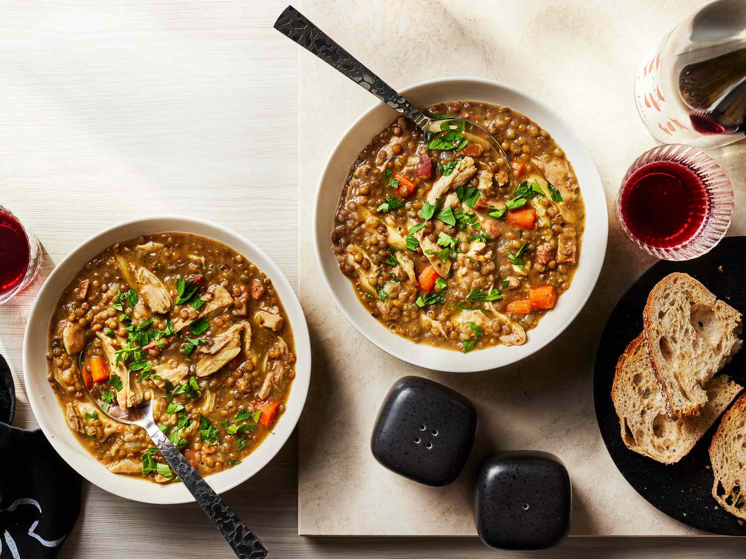 2 bowls Chicken Lentil stew with a plates of sliced bread on the side, black salt and pepper shakers, and a glass and bottle of red wine 