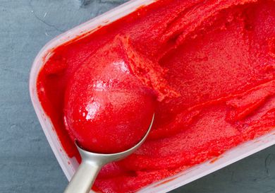 A rectangular container of bright red strawberry sorbet with a scoop being pulled from it.