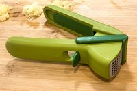A garlic press placed on a wooden surface with some minced garlic around it
