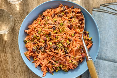 Carrot slaw in a blue bowl surrounded by glasses of water on a wooden surface with napkins and serving spoon 