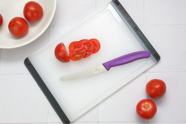 A serrated paring knife with a purple handle slicing tomatoes on a cutting board with additional tomatoes nearby