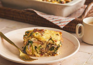 Serving of baked casserole square from the side on a plate with a gold fork. Coffee and basket of bigger casserole are in the background. 