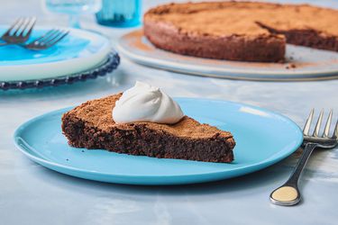 Slice of flourless chocolate cake, topped with a dollup of cream, on a blue plate, blue tabletop, and the cake is in the background 