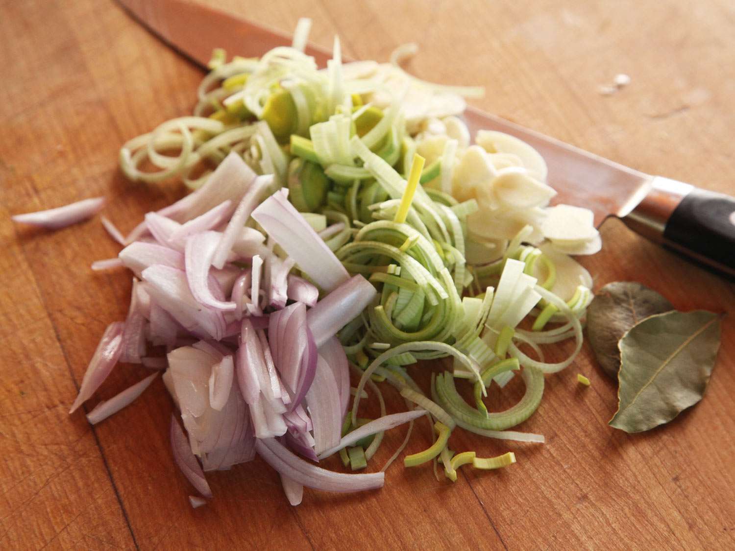 Sliced shallot, leek, and garlic mounded on a cutting board next to a pair of bay leaves.