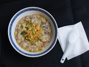 Overhead view of a bowl of brown rice congee with beef, shiitake, and garlic chips, served on a black table with a soup spoon.