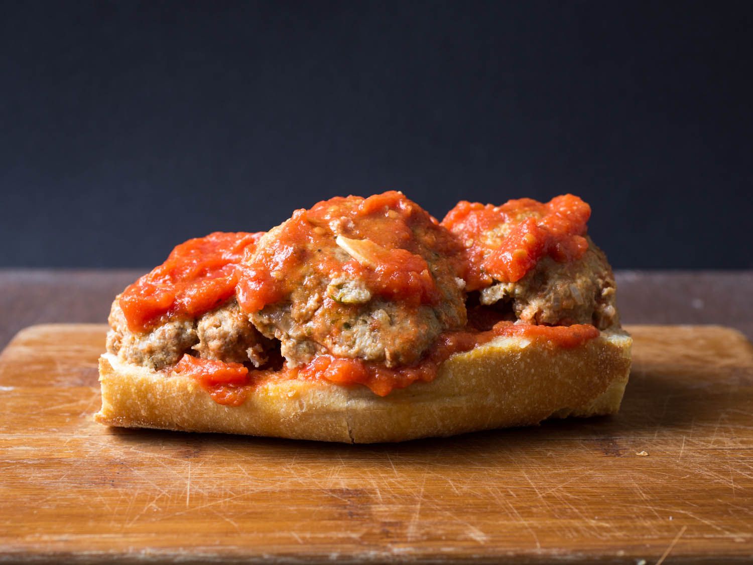 Profile view of the meatball sandwich being constructed. The bottom of the Italian bread has been coated with sauce, layered with halved meatballs, followed by more sauce.
