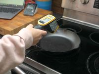 A person measuring the surface temperature of a cast iron skillet on a stovetop with an infrared thermometer