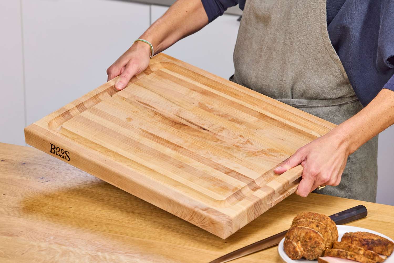 Person holding a John Boos Reversible Maple Wood Cutting Board with Juice Groove over a table with food and a knife