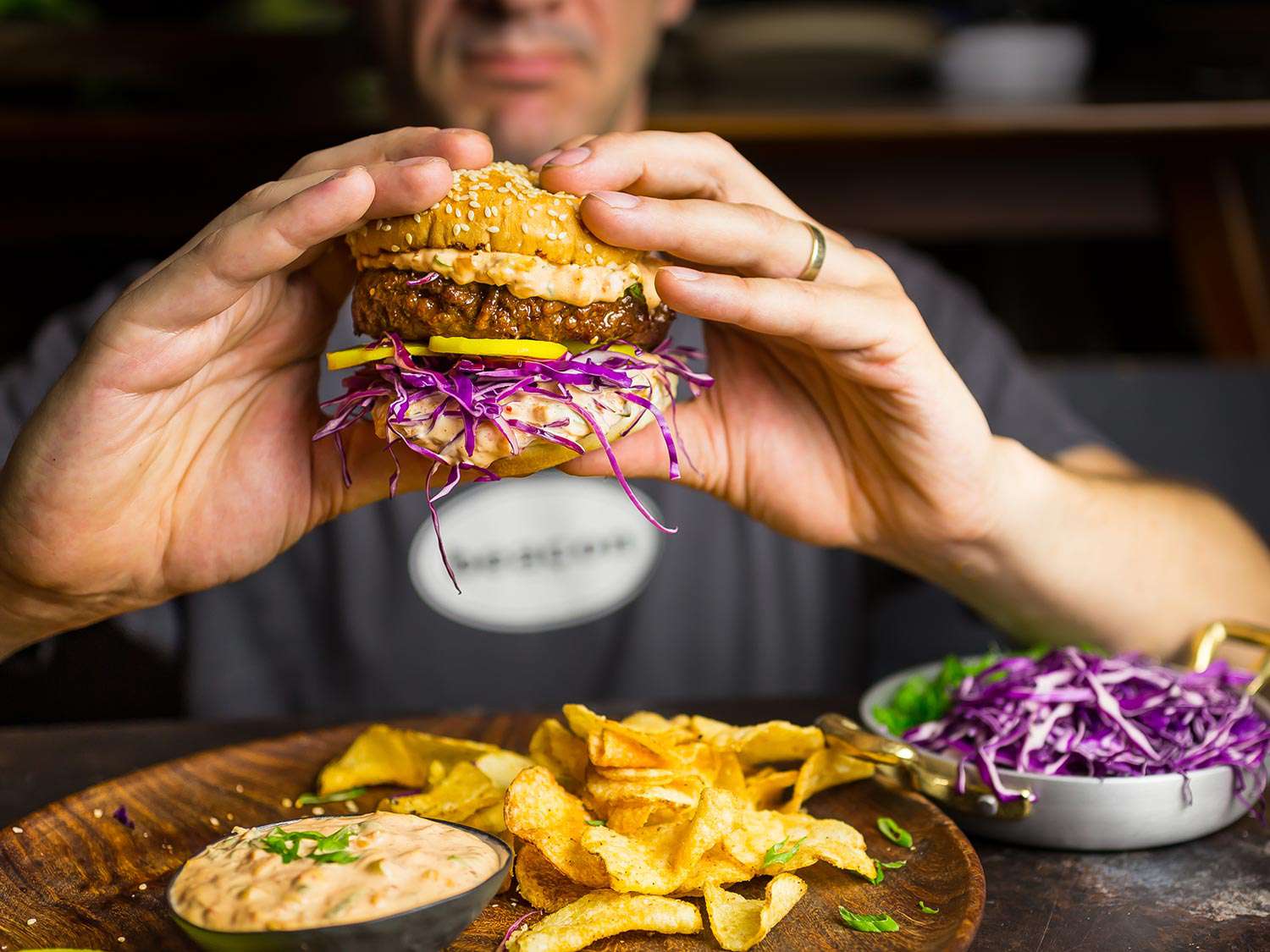 A hand holding a bulgogi burger over a serving platter of chips and kimchi mayonnaise, with a plate of red cabbage on the side. 