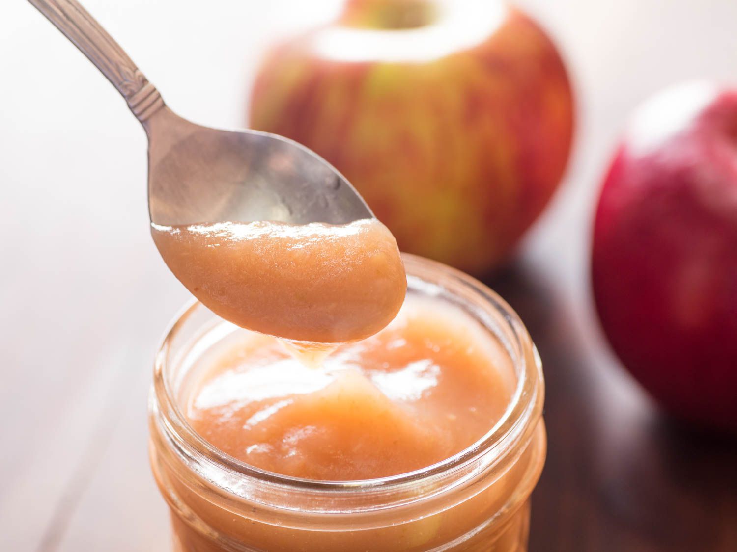 Close-up of a spoon lifting some very smooth applesauce out of a jar, with whole apples in the background.
