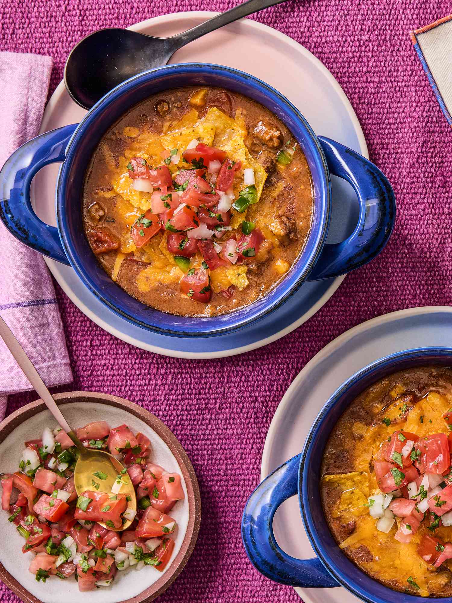 Overhead of 2 blue bowls with taco soup, on with a spoon in it, a small dish with pico de gallo on it, and a magenta colored textile surface. 